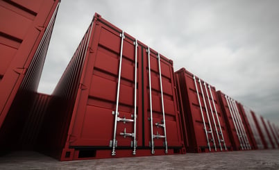 A row of red shipping containers with metal locks and handles lined up on a concrete surface. The perspective is low and angled, showing the containers stretching into the distance under a cloudy sky.