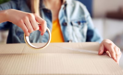 A person sealing a cardboard box with clear packing tape. The individual is wearing a denim jacket and holding the tape in one hand while applying it to the box.