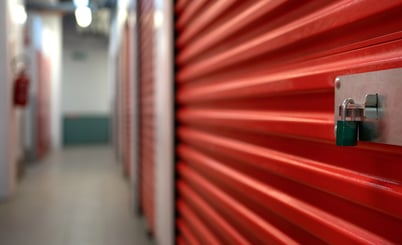 A row of red storage unit doors in a hallway, with a padlock securing one of the doors on the right. The corridor is well-lit, and the doors are metallic with horizontal grooves. The focus is on the foreground door, while the background appears slightly blurred.