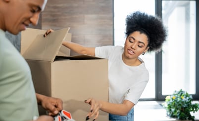 A woman wearing a white shirt is packing items into a large cardboard box while another person in a blurred green shirt is holding tape. They are in a brightly lit room with large windows and a plant in the background.